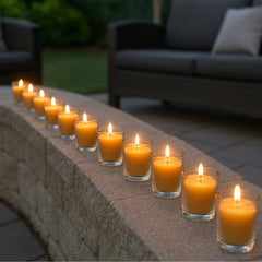 Twelve lit beeswax votive candles in clear glass holders arranged in a curved line on a stone retaining wall in an outdoor patio setting at twilight.