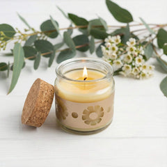 Beeswax candle in a glass jar with cork lid on a white surface with greenery and flowers.