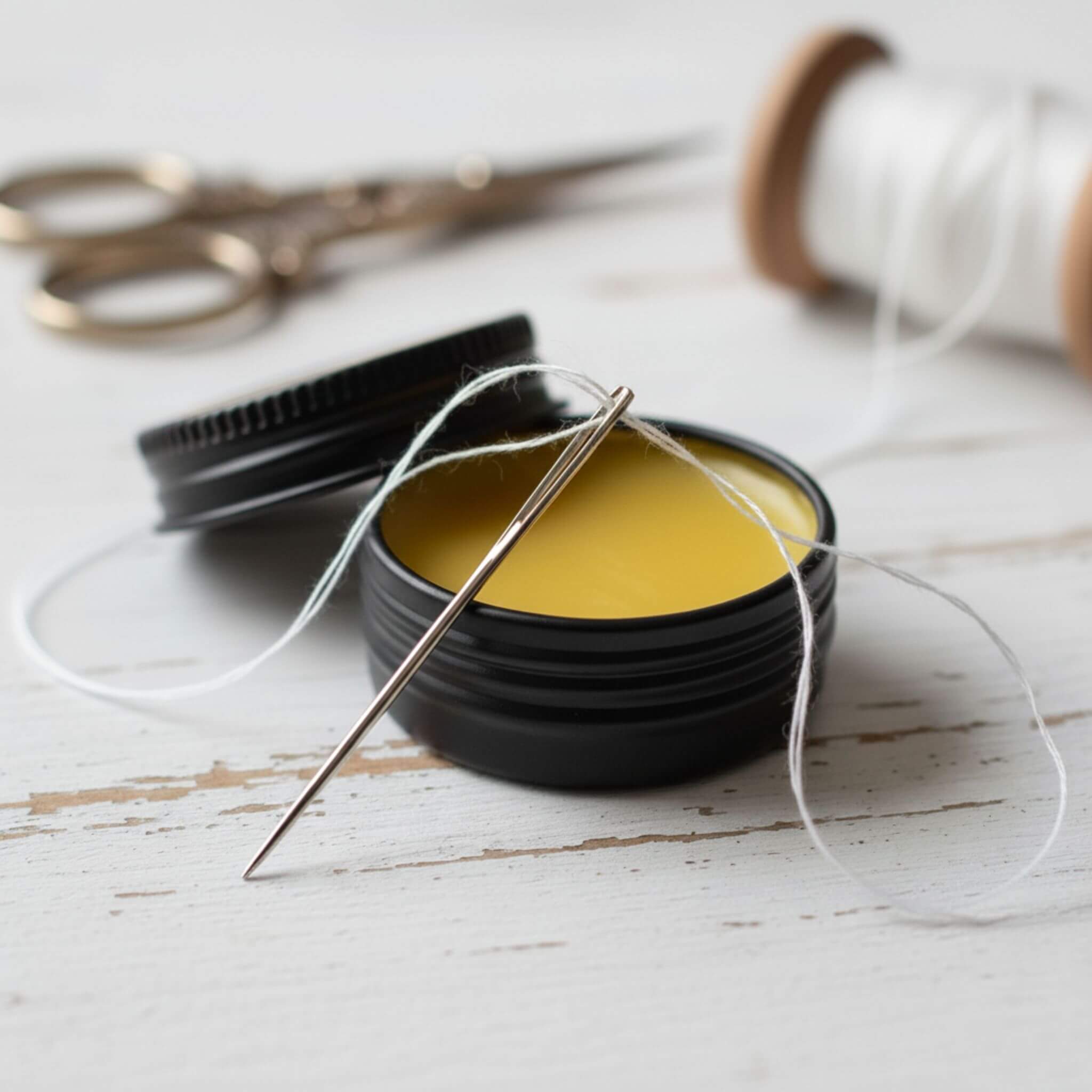 Blossom & Hive Beeswax non-removable beeswax thread wax in a black tin, a needle, and scissors on a white wooden surface.