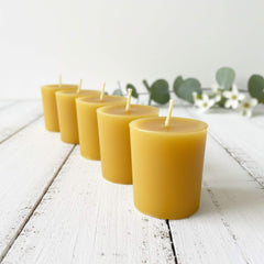 Blossom & Hive 6 beeswax votive refills lined up in a row, on a white wooden surface with greenery in the background