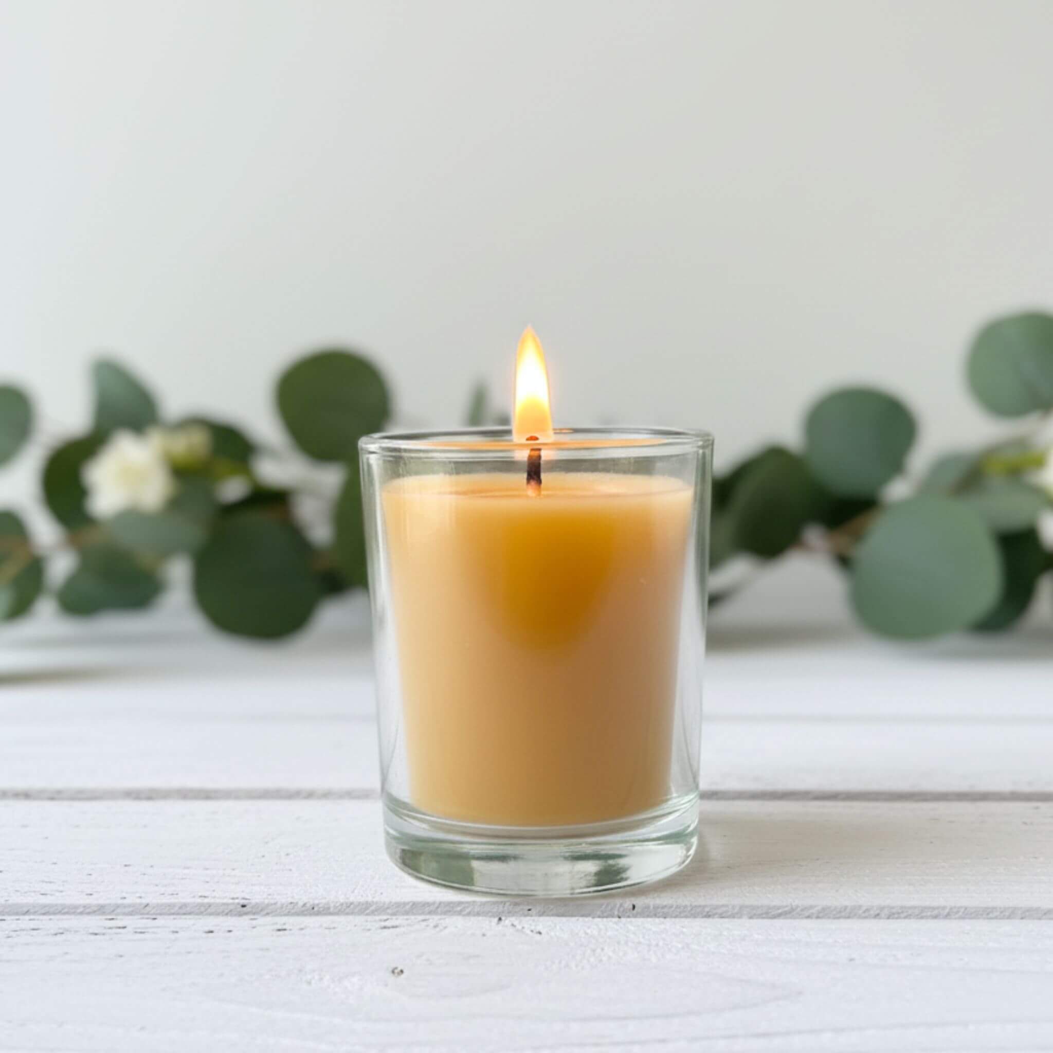 A lit Blossom and Hive beeswax votive in a glass holder with a white background and greenery