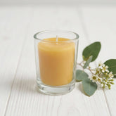 Beeswax votive in a glass holder with a small sprig of greenery beside on a white painted timber surface