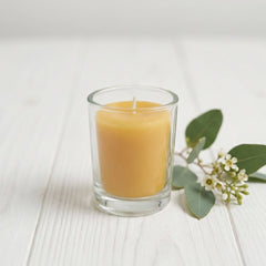 Beeswax votive in a glass holder with a small sprig of greenery beside on a white painted timber surface
