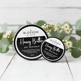 Blossom and Hive Honey Butter Balm for dry skin, available in 2 sizes, two black tins sitting on a white timber surface with blurred greenery in the background