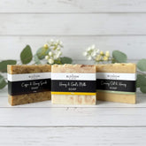 Three bars of cold-processed, handmade Blossom and Hive honey soap with labels on a wooden surface with greenery in the background