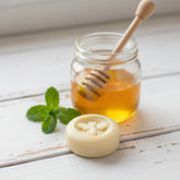 A Blossom and Hive Honey and Mint lotion bar, a jar of honey with a wooden dipper, and mint leaves on a wooden surface.
