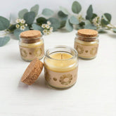 Three small jars with cork lids containing beeswax candles on a white surface with greenery in the background.
