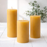 Three yellow beeswax pillar candles on a white surface with a blurred plant in the background