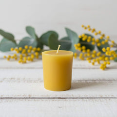 Blossom & Hive single Beeswax votive on a wooden surface with green leaves and yellow flowers in the background