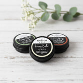 Blossom and Hive lip balm and lip scrub trio in black aluminium tins, sitting on a white painted timber surface with blurred greenery in the background