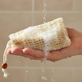 Hand holding an eco-friendly cotton soap saver bag with water running over it against a tiled wall background