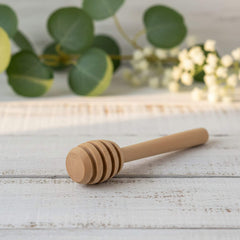 Wooden honey dipper on a light wooden surface with green leaves and lights in the background
