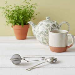 Blossom & Hive stainless steel mesh tea ball and heart-shaped strainer on a white timber surface with a teapot, a tea cup and a pot plant in the background