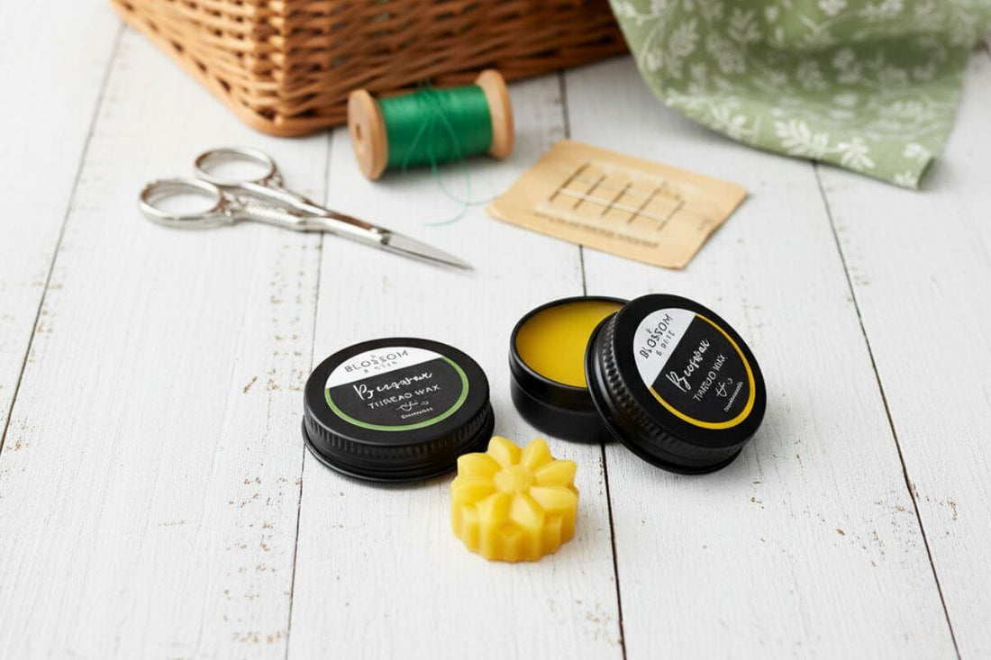 Two varieties of beeswax thread wax in black tins alongside sewing accessories on a white painted timber surface