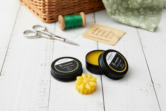 Two varieties of beeswax thread wax in black tins alongside sewing accessories on a white painted timber surface