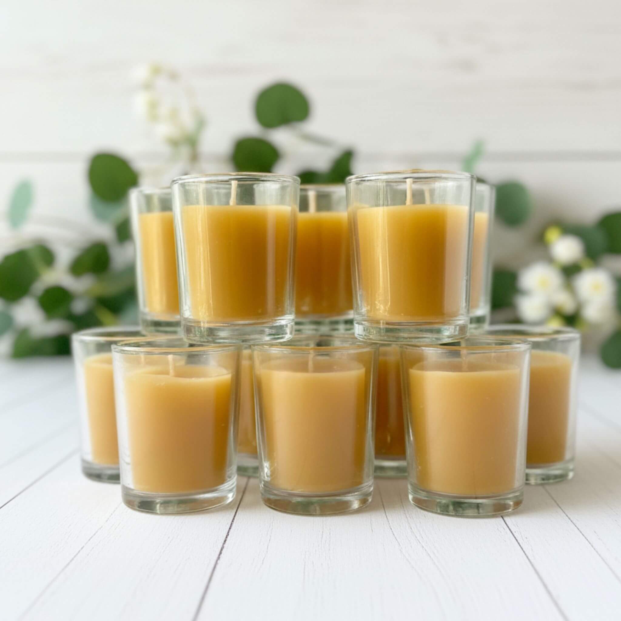 Blossom and Hive beeswax votives in glass candle holders  - 12 pack, placed on a white background with blurred greenery at the back