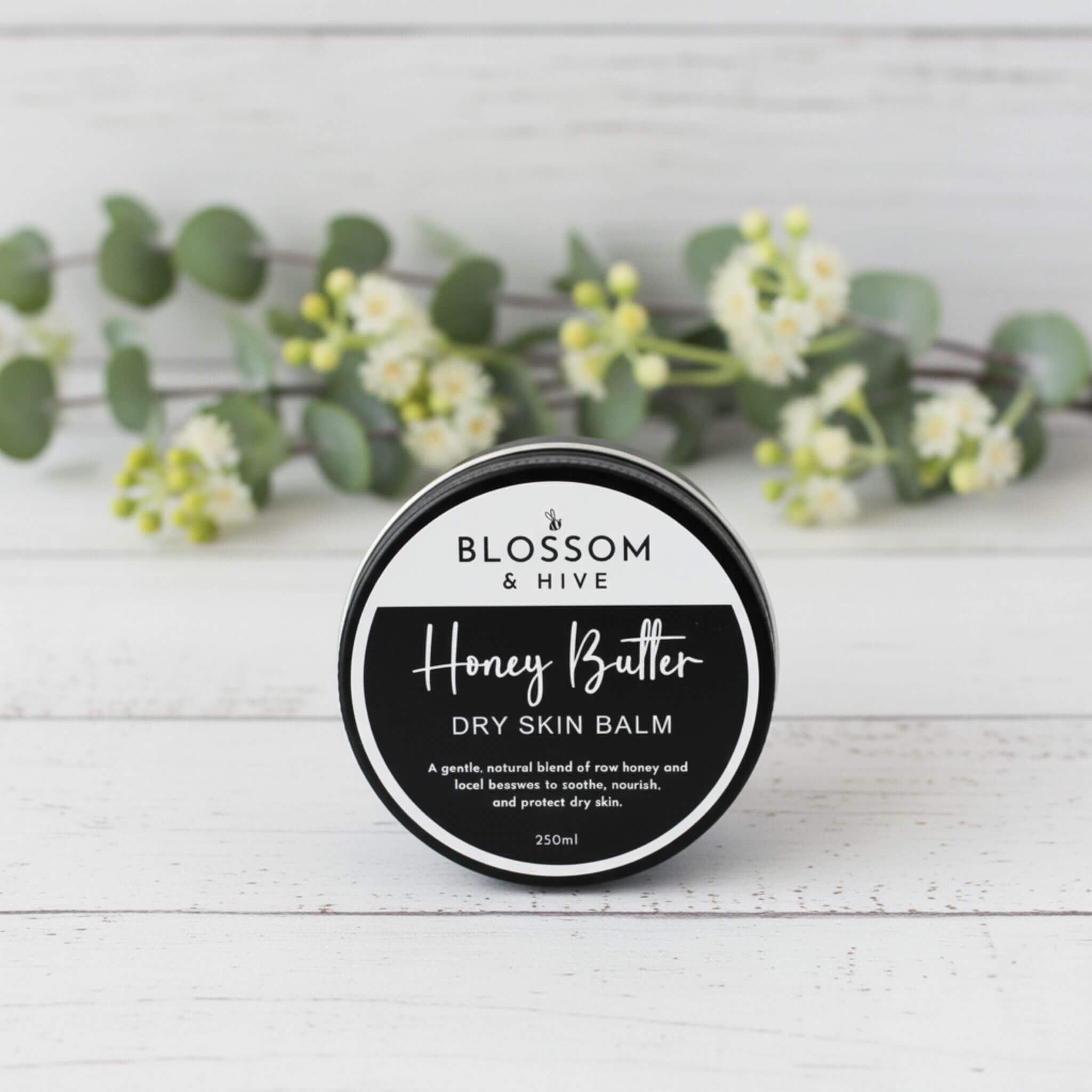Blossom and Hive Honey Butter Balm 250ml for dry skin, in a black tin sitting on a white timber surface with blurred greenery in the background