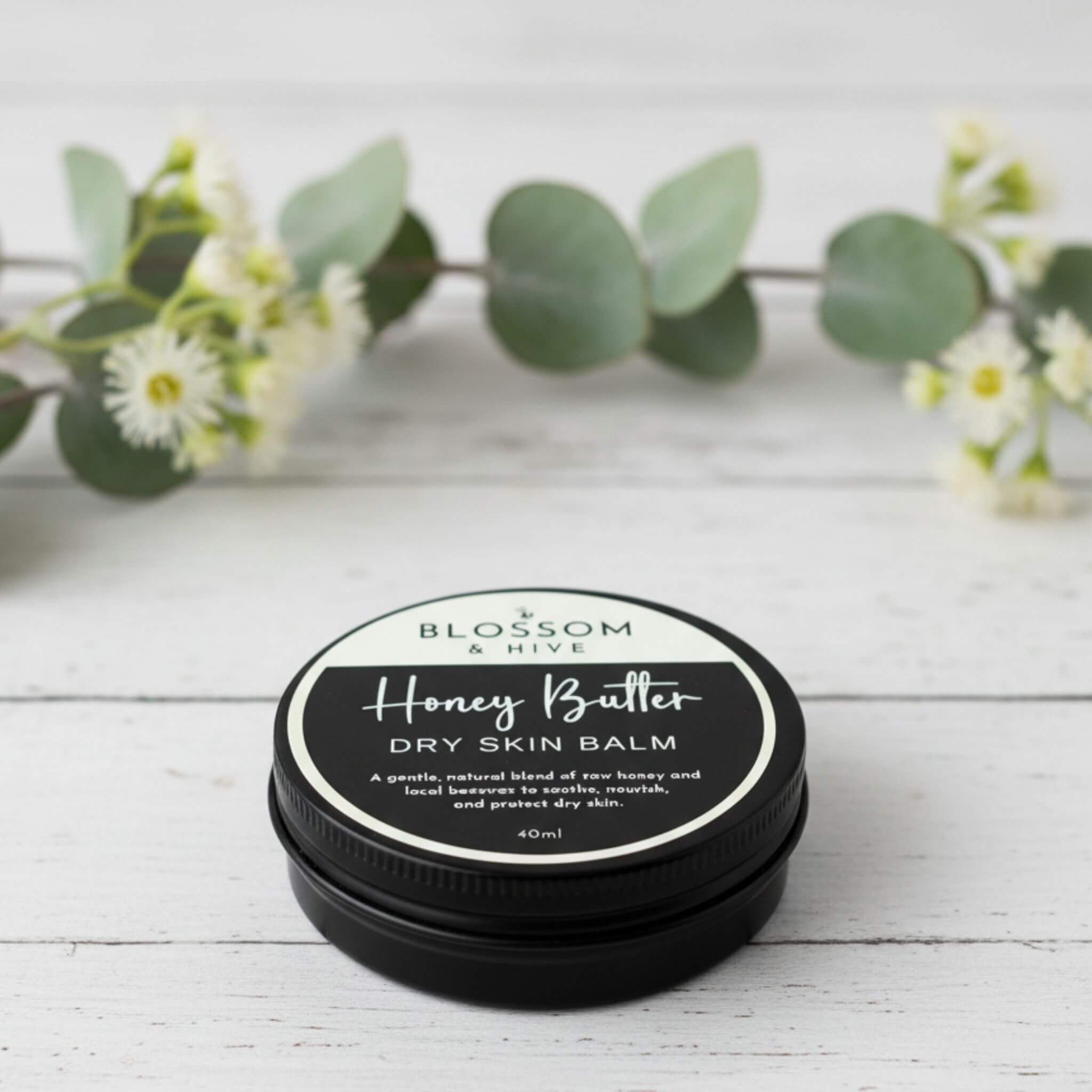 Blossom and Hive Honey Butter Balm 40ml for dry skin, in a black tin sitting on a white timber surface with blurred greenery in the background