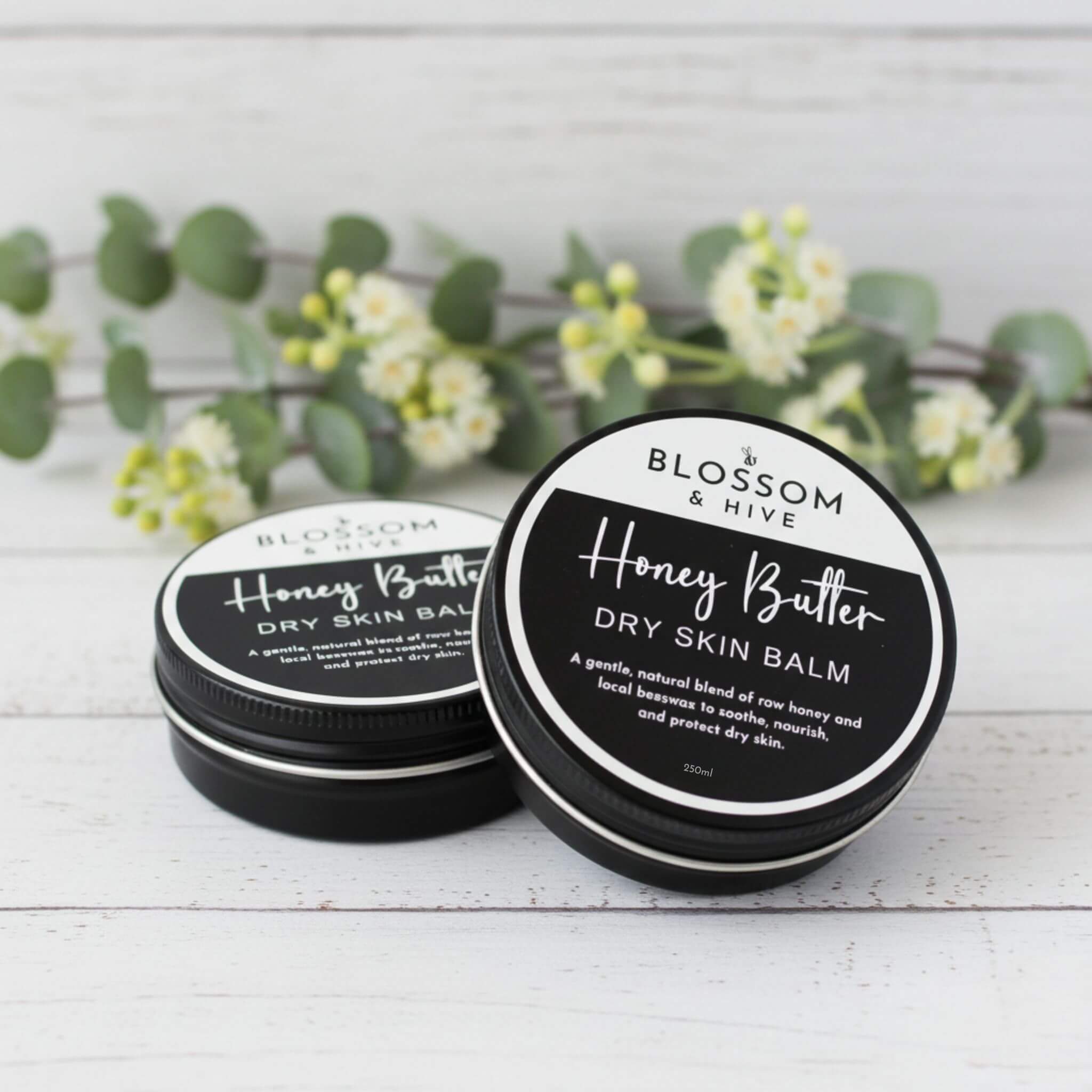 Blossom and Hive Honey Butter Balm for dry skin, 2x250ml black tins sitting on a white timber surface with blurred greenery in the background
