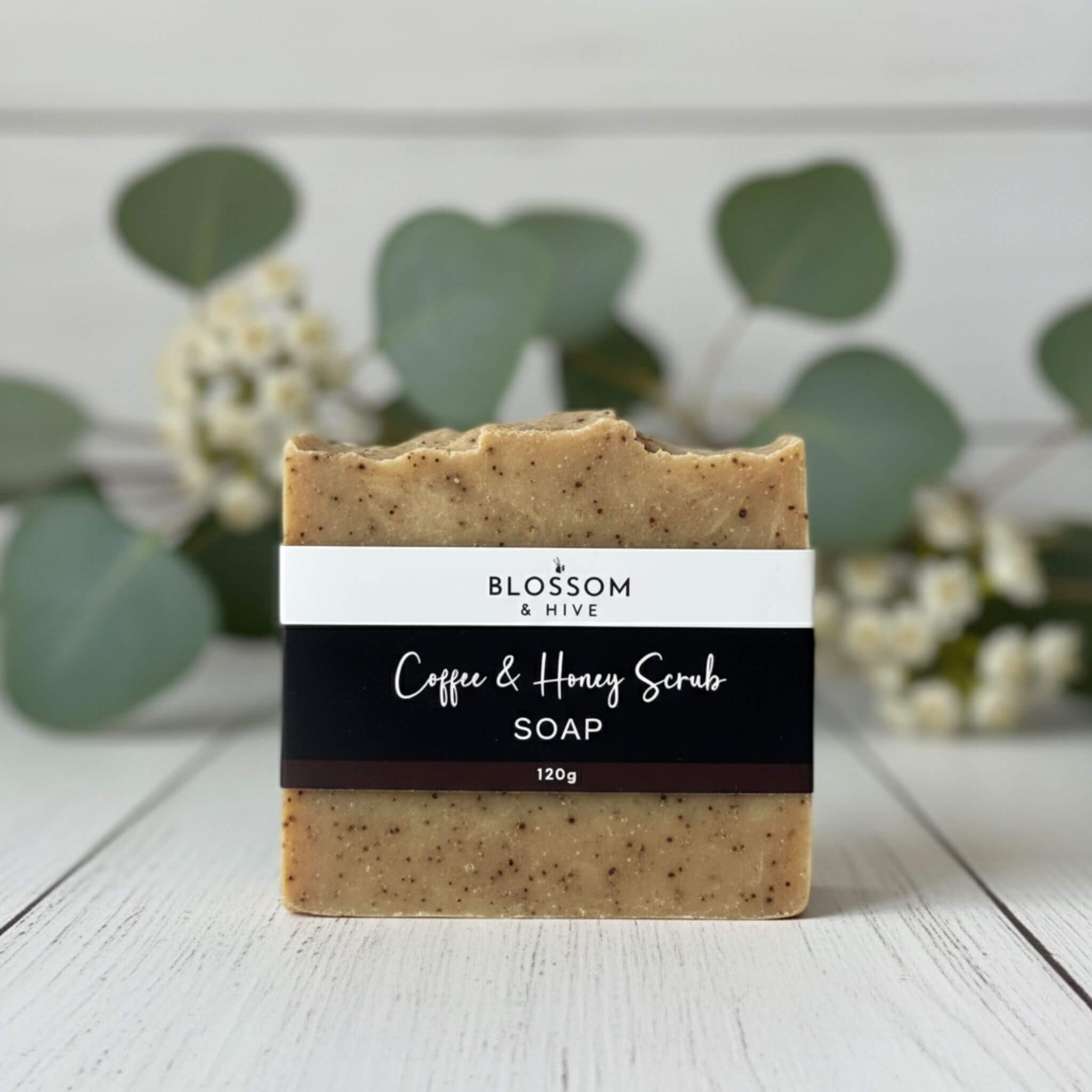 Bar of Blossom and Hive handcrafted coffee and honey scrub soap on a wooden surface with greenery in the background