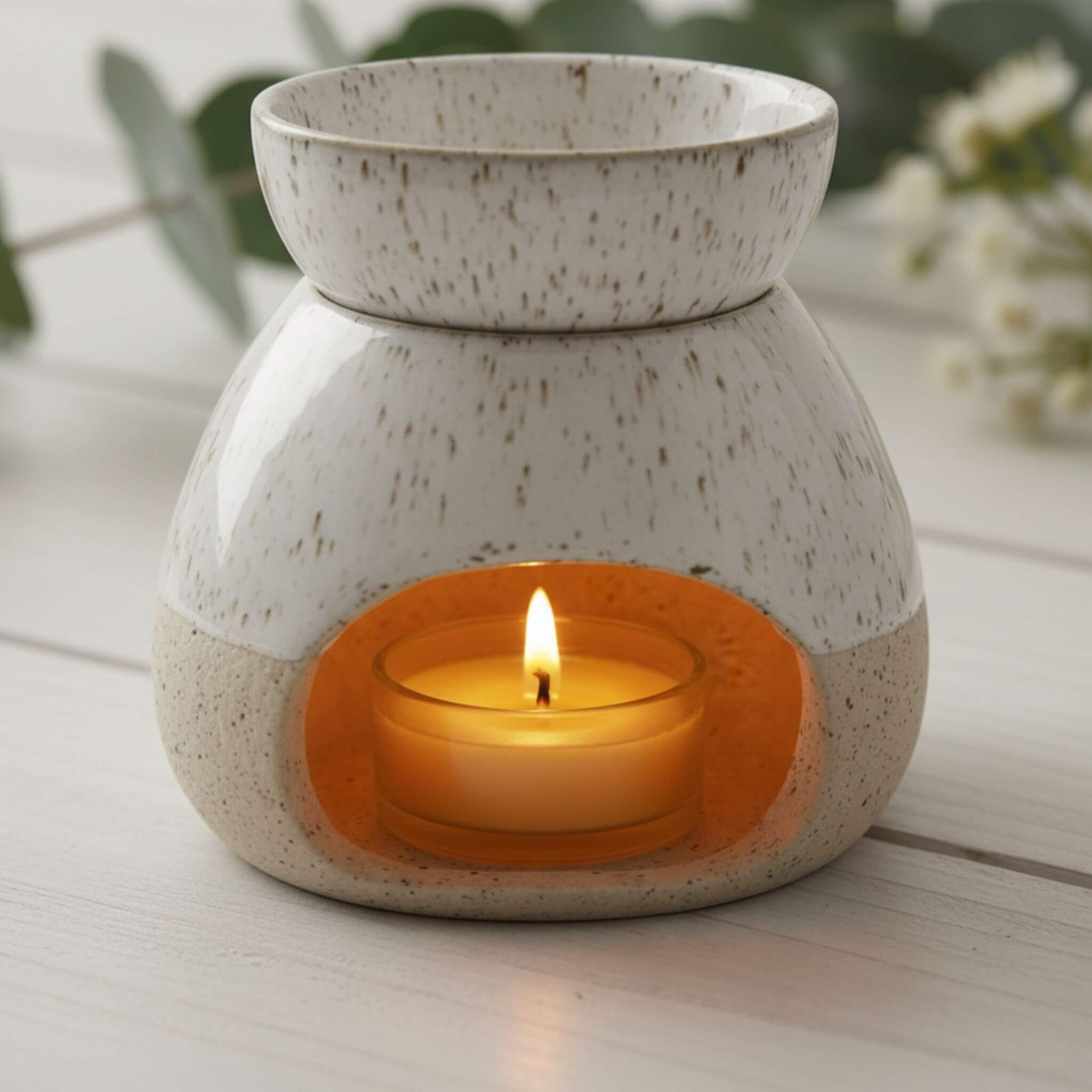 Speckled ceramic oil burner with a lit Blossom and Hive beeswax tealight  on a wooden surface.