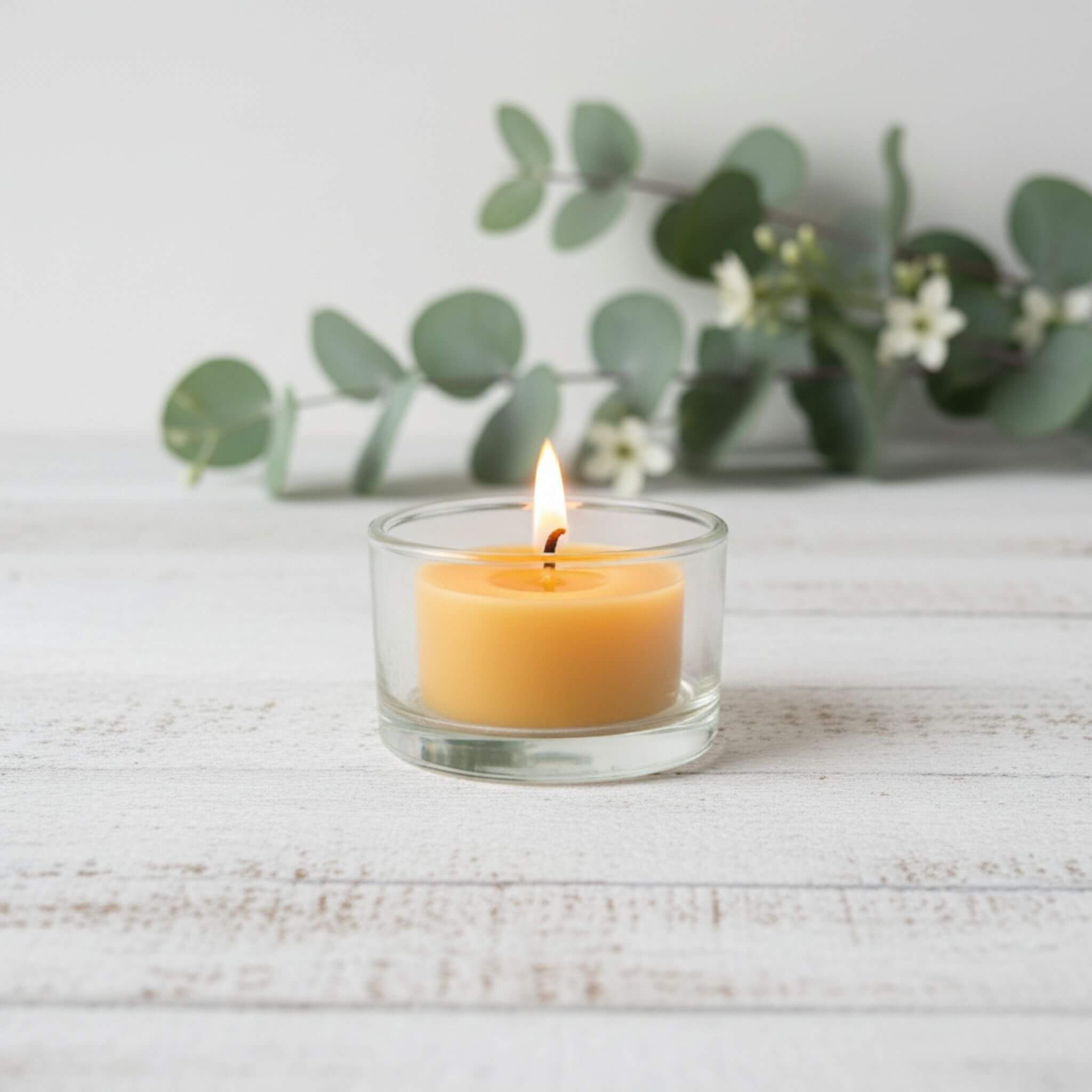 Unscented beeswax tealight candle in a clear glass holder on a light wooden surface with greenery in the background.