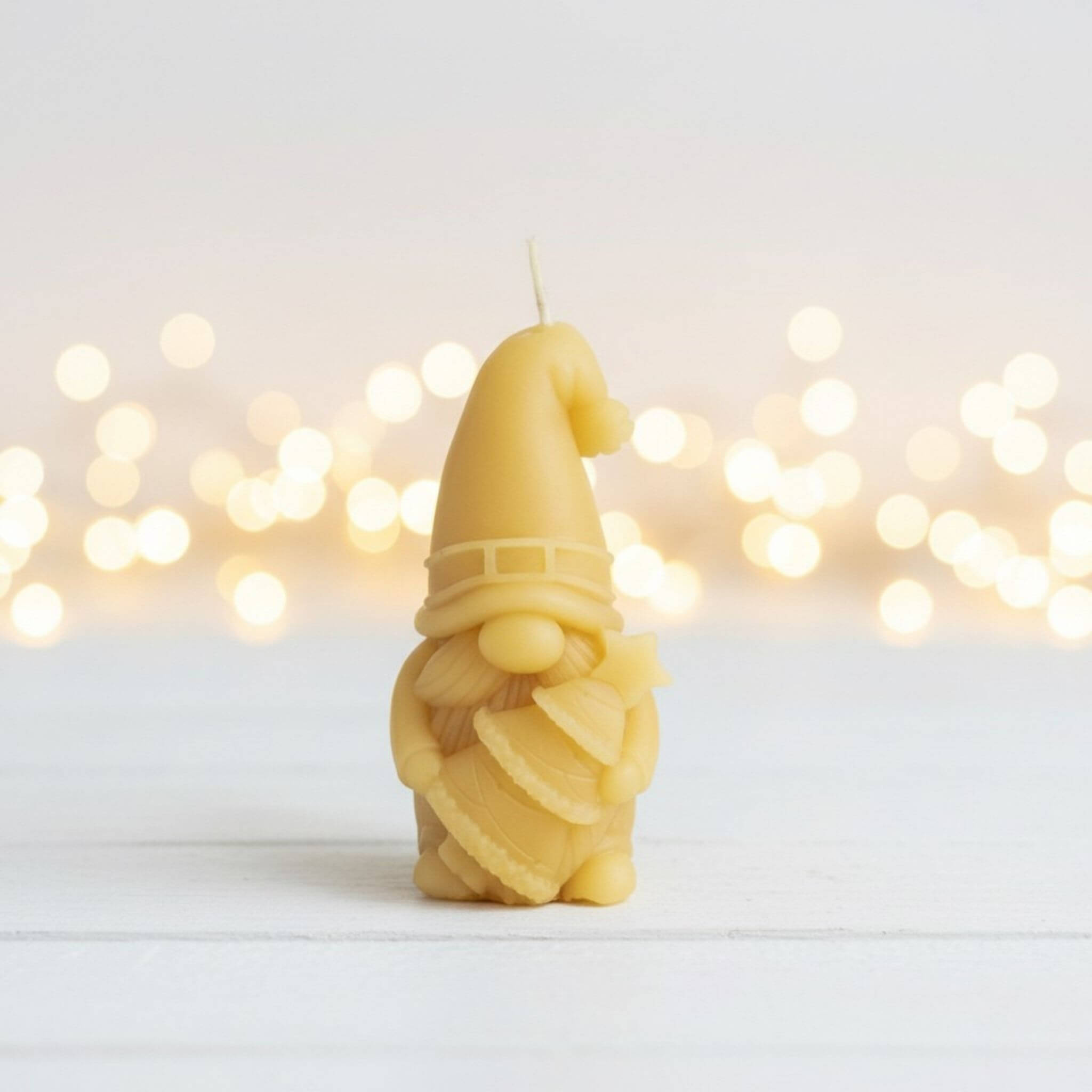 Blossom and hive beeswax candle shaped like a gnome and holding a christmas tree on a white surface with blurred lights in the background