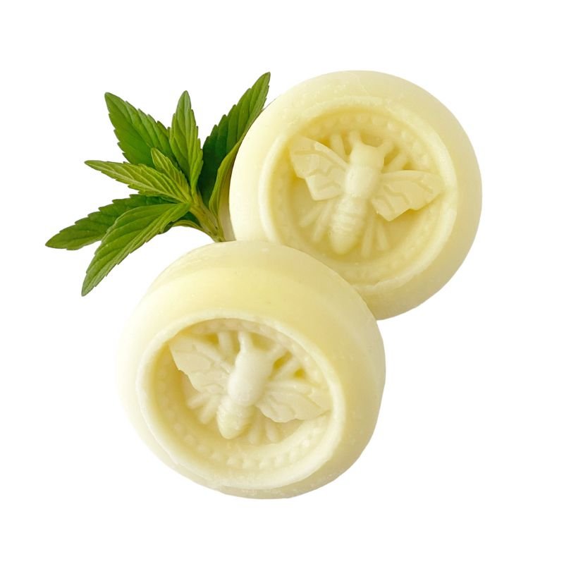 Two round honey and mint lotion bars embossed with a bee design, placed against a white background with fresh mint leaves. A natural skincare product for dry heels and elbows.
