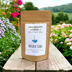 A brown kraft pouch of Collombatti Naturals Masala Chai tea, standing on a rustic wooden surface with a lush countryside and blooming flowers in the background. The label features a minimalist blue teacup illustration and highlights the brand’s commitment to ethical, sustainable, and natural products.